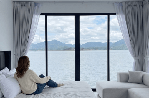 Cortina gris de paletón francés instalada en un dormitorio moderno con gran ventanal y vista hacia un río, diseño elegante de Cortinas Home Doral.
